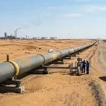 An industrial pipeline stretches across a vast desert landscape under a clear blue sky. Two workers in blue uniforms and safety gear inspect a valve station next to the pipeline, where a white SUV is parked on a dirt path. In the background, sand dunes, electrical pylons, and a distant oil refinery with smokestacks are visible.