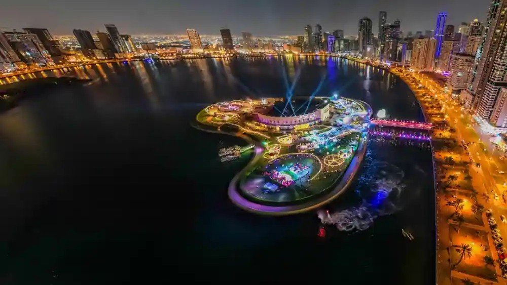 Aerial night view of Al Majaz Waterfront in Sharjah with illuminated skyline, Khalid Lagoon, and colorful waterfront attractions.