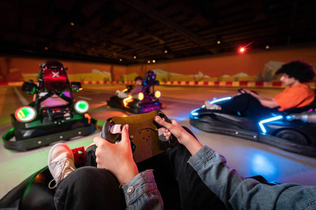 A first person view of visitors driving illuminated electric karts at Chaos Karts Dubai while navigating an indoor augmented reality racing arena with glowing tracks and interactive gaming effects.