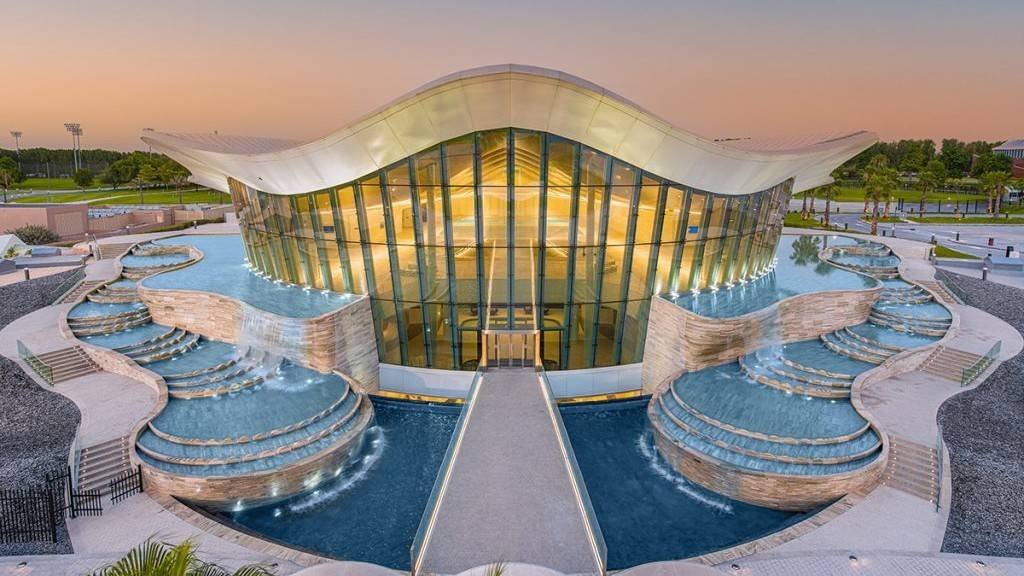 Deep Dive Dubai exterior building in Nad Al Sheba with modern curved architecture, glass façade, cascading water features and entrance bridge at sunset.