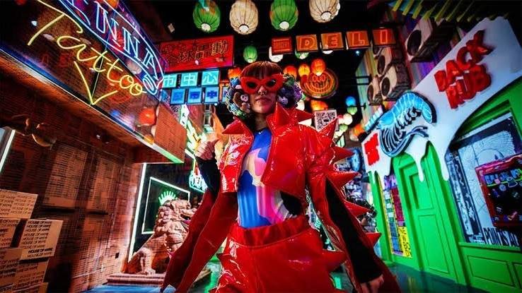 A visitor wearing a bright red futuristic outfit and sunglasses walks through a neon lit interactive room at House of Hype Dubai, surrounded by colourful signs, lanterns, digital displays, and immersive themed attractions.