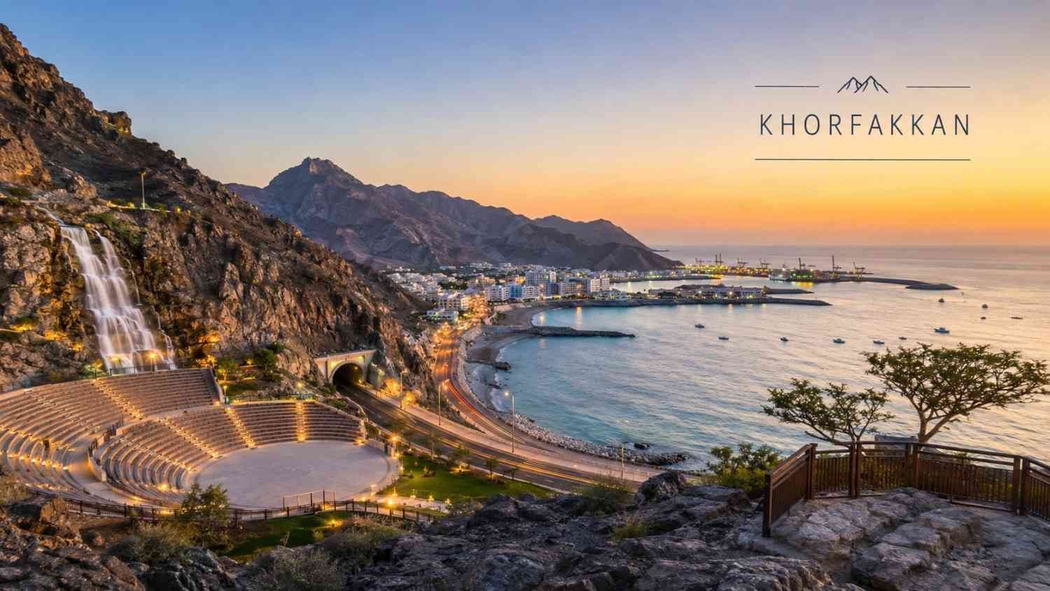 Khorfakkan Amphitheatre and artificial waterfall at sunset, overlooking the Gulf of Oman with rugged Hajar Mountains, coastal road, harbor, and waterfront illuminated by evening lights in Sharjah, UAE.