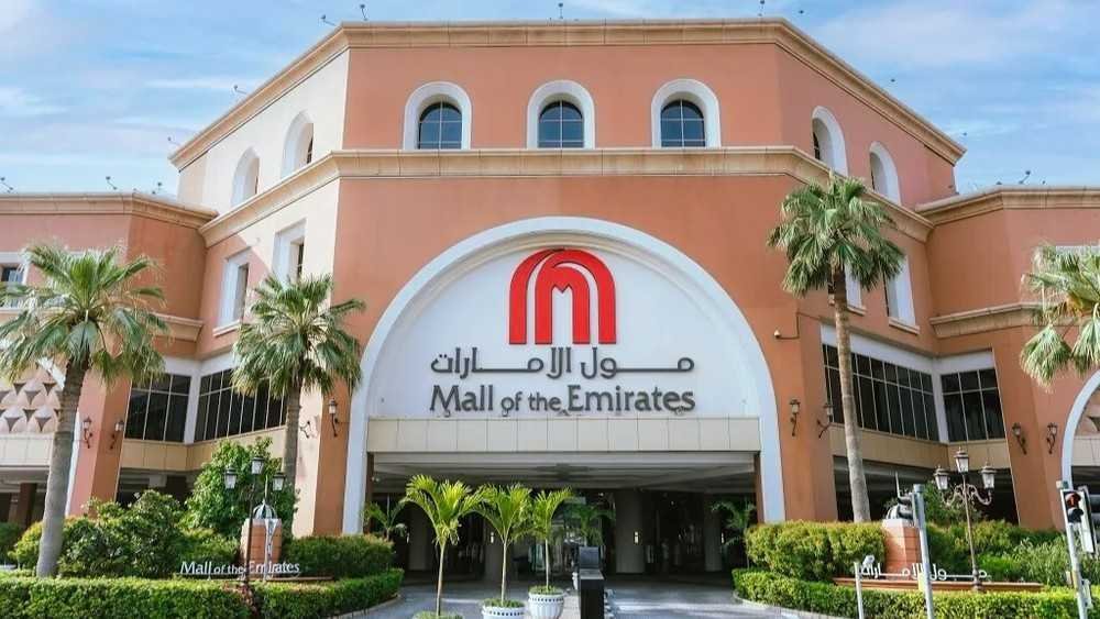 Mall of the Emirates main entrance in Al Barsha, Dubai, with the iconic red logo, palm trees, landscaped gardens and the grand exterior welcoming visitors.