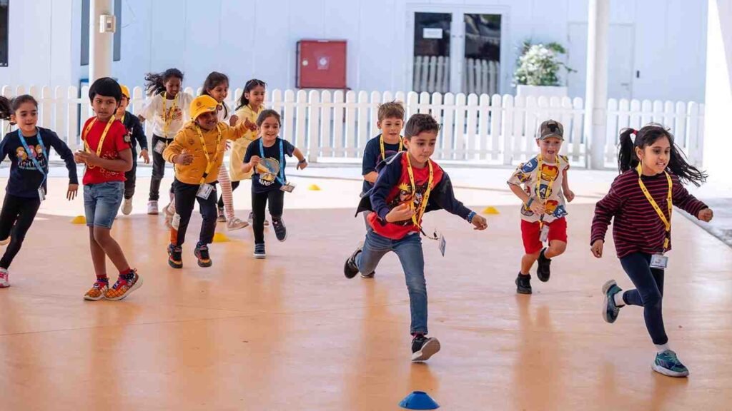 Children participating in an indoor UAE summer camp activity, running through a supervised obstacle course while wearing name badges in a bright, air conditioned learning environment.