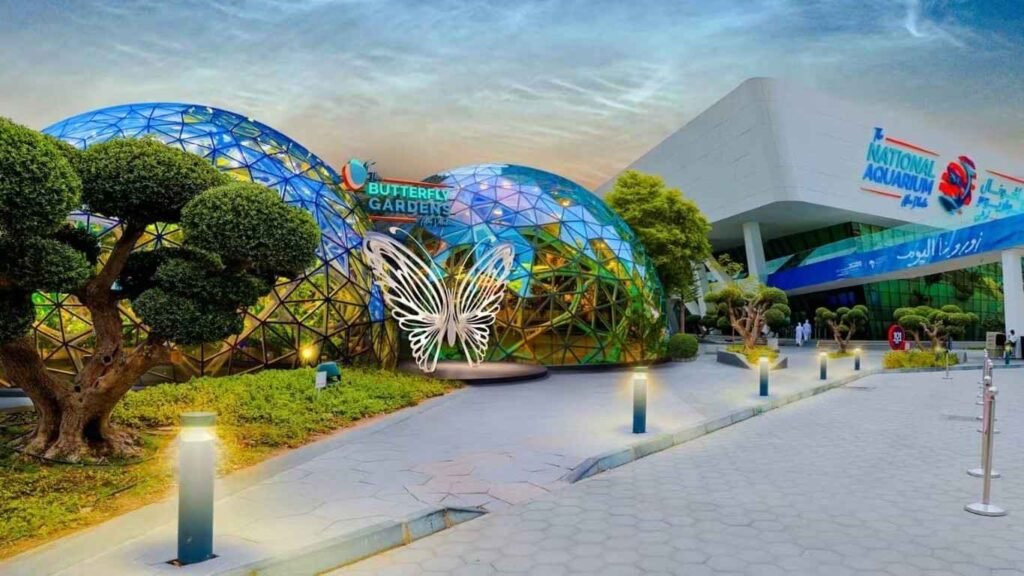 Exterior view of The Butterfly Gardens Abu Dhabi at Al Qana, featuring two glass domes with a butterfly entrance sculpture beside The National Aquarium building.