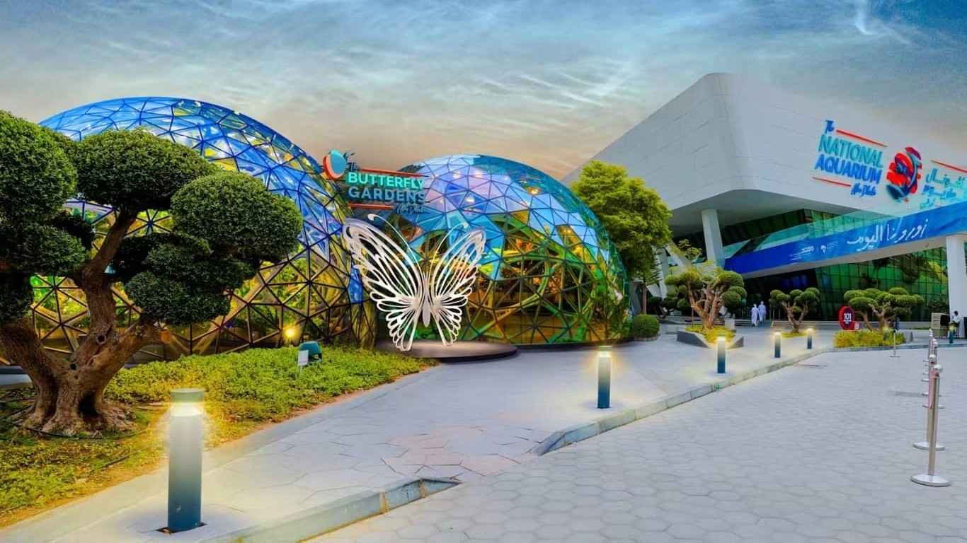 Exterior view of The Butterfly Gardens Abu Dhabi at Al Qana, featuring two glass domes with a butterfly entrance sculpture beside The National Aquarium building.