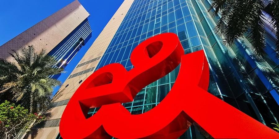 Close up of the e& logo in front of the company's glass headquarters building with modern skyscrapers and palm trees under a clear blue sky.