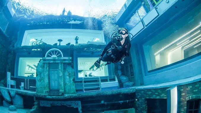Scuba diver exploring the underwater Sunken City at Deep Dive Dubai, featuring submerged buildings, large viewing windows and themed dive attractions inside the world's deepest diving pool.