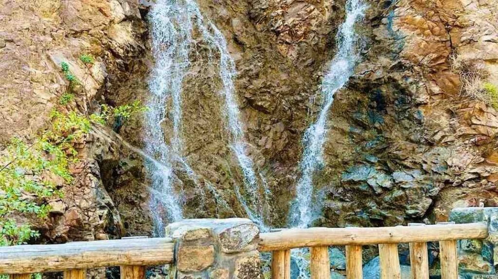 Close up of the 25 metre waterfall at Shees Park Khorfakkan flowing over rocky cliffs, with a wooden viewing fence in the foreground.