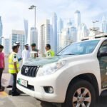Dubai Police officers and traffic officials stand beside a white patrol SUV during a roadside inspection in Dubai, with several vehicles and the city's skyline visible in the background.