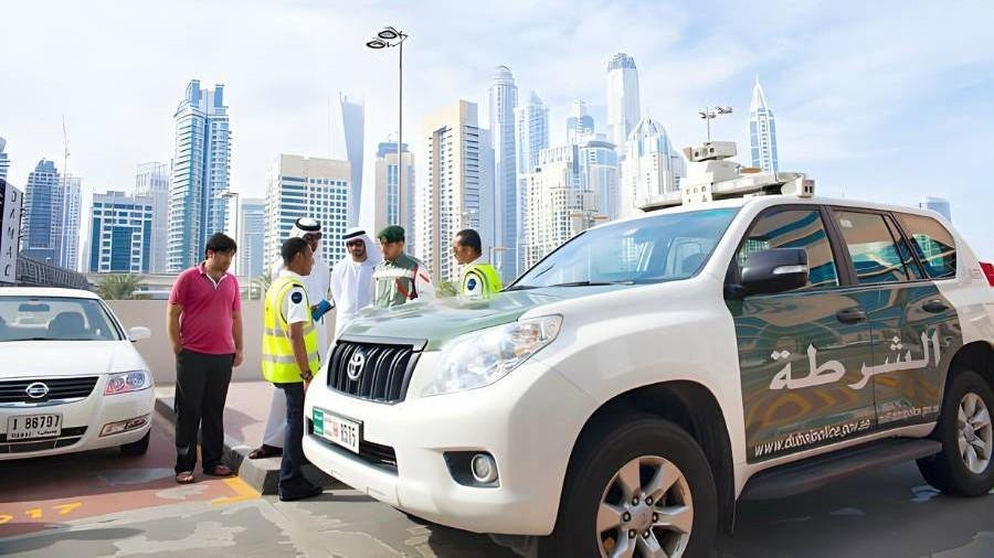 Dubai Police officers and traffic officials stand beside a white patrol SUV during a roadside inspection in Dubai, with several vehicles and the city's skyline visible in the background.