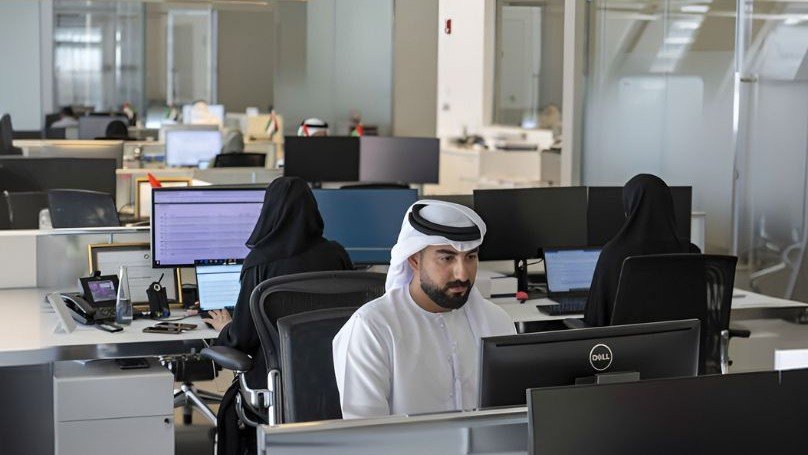 Employees work at computer stations inside a modern Dubai office, with staff in traditional Emirati attire and an open plan workspace featuring multiple desks, monitors, and glass partitions.