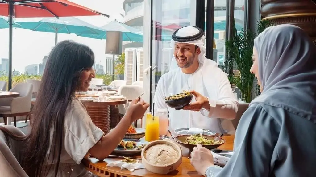 Customers enjoying a meal at a restaurant in the UAE as a server presents a dish, highlighting the welcoming dining experience and social atmosphere during the summer season