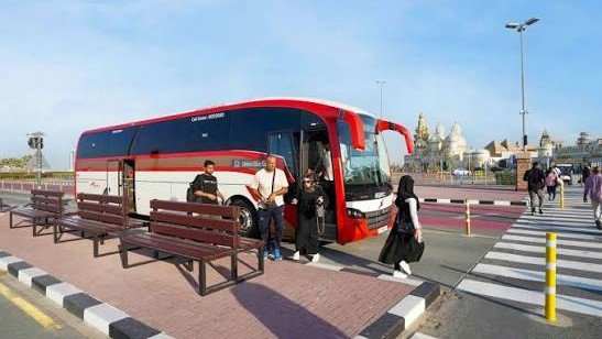 Passengers board a red Dubai public transport bus at a roadside stop near a pedestrian crossing, with city landmarks visible in the background.